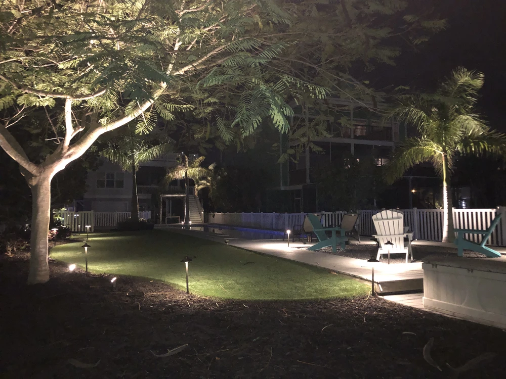 Landscape lighting highlights a broad tree and curved green lawn beside a pool deck with white and teal chairs.